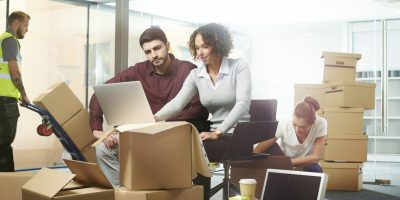 A business owner sits amongst his moving in boxes , waiting for the removal team to bring the desks . An interior designer is showing him the modifications needed as building contractor brings in various fixtures and fittings for the new office layout . They are looking at the layout plans and computer drawings . In the background her colleague is checking the files.